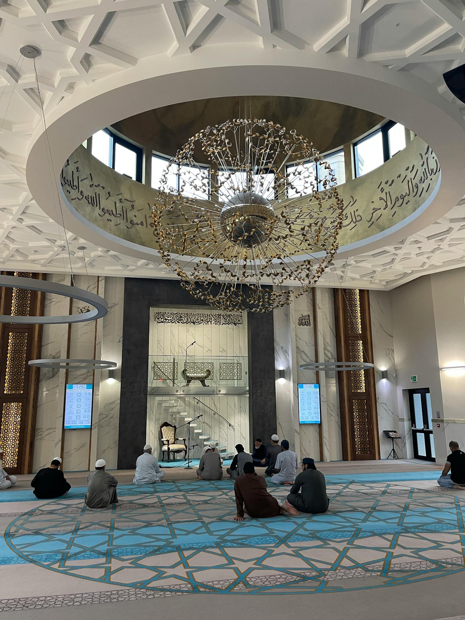 Group of people in prayer inside a beautifully designed mosque with intricate ceiling chandelier.